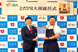 Zwei lächelnde Männer mit einem Zertifikat, mit einem Banner im Hintergrund, das 'Toda City Wins PR Award' lautet und ein Foto einer Person zeigt.