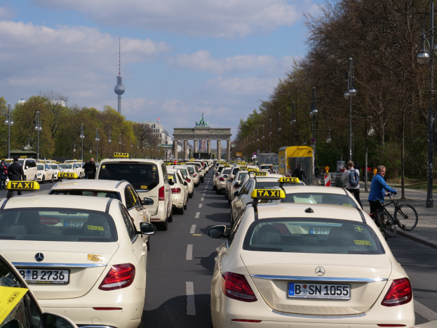 Eine lange Reihe von Taxis, die an einer belebten Straße in Berlin, Deutschland, geparkt sind, mit Fahrradfahrern und Fußgängern auf dem Gehweg, flankiert von Bäumen und Laternenmasten und Gebäuden, einem Bogen und einem Turm im Hintergrund unter einem bewölkten Himmel.