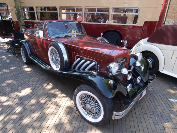 Ein Vintage-Auto neben einem Doppeldeckerbus auf einem Kopfsteinpflaster, mit Stühlen und einem Baum im Vordergrund, einem Gebäude mit Fenstern im Hintergrund und Menschen im Bus zu sehen.
