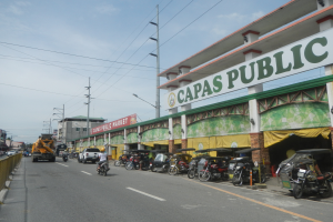 Eine belebte Stadtstraße mit Fahrzeugen, einem Geländer auf der linken Seite, Gebäuden, Polen, Laternen, Schildern und einigen Menschen unter einem bewölkten Himmel, mit einem Gebäude auf der Seite, auf dem eine Tafel mit der Aufschrift 'Capas Public' zu sehen ist.