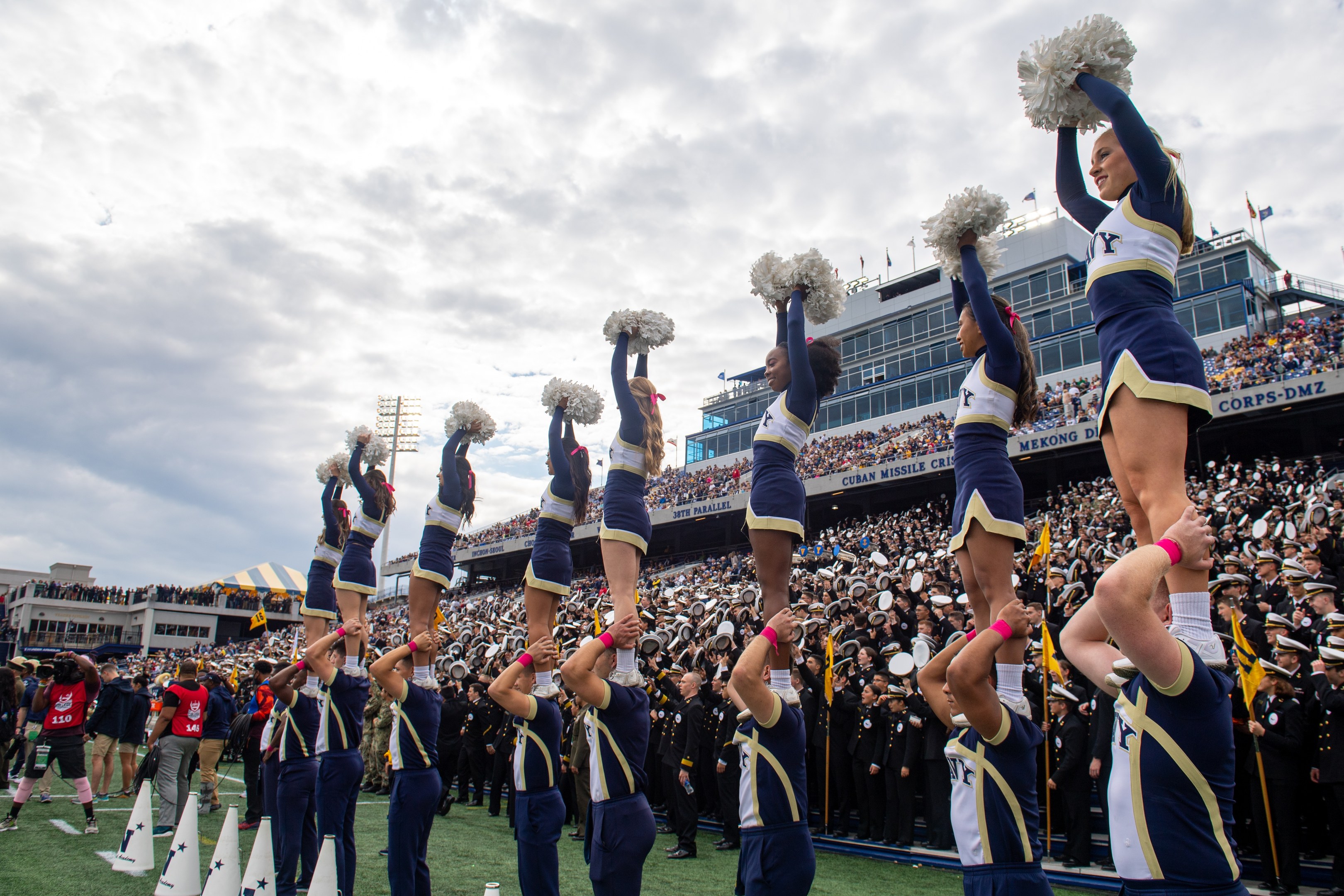 Eine Gruppe von Cheerleadern in blauen und weißen Uniformen führt einen Stunt auf einem Stadionevent durch, während sie Pompons halten und eine Person die Szene filmt.