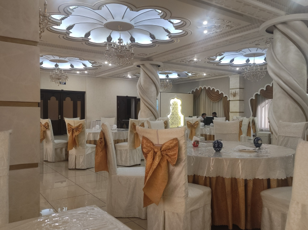 Ein großer Festsaal mit Tischen und Stühlen, die mit Tüchern drapiert und mit verschiedenen Gegenständen geschmückt sind, Wände mit Türen und Vorhängen und eine Decke mit Lichtern und Kronleuchtern.
