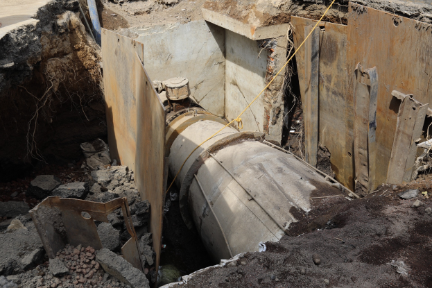 Ein großes Metall-Rohr für eine Klärgrube sitzt in einem Schmutzfeld umgeben von Steinen, Schlamm und anderen Gegenständen, mit einer Straße auf der linken Seite.