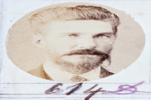 Ein altes Foto eines Mannes mit Bart und Schnurrbart, der direkt in die Kamera schaut und dessen Lippen leicht geöffnet sind.