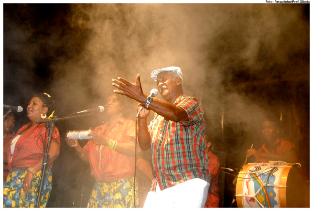 Eine Gruppe von Musikern, die auf der Bühne stehen, mit einem Mann, der in ein Mikrofon singt, in der Mitte und einem anderen Mann, der rechts ein Schlagzeug spielt.