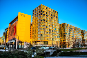 Eine belebte Stadtlandschaft mit hohen Gebäuden, darunter das Jurys Inn Birmingham City Centre, mit Fahrzeugen auf der Straße, Straßenlaternen, Straßenbeleuchtung, Verkehrsampeln, Büschen, Bäumen und einem klaren blauen Himmel.