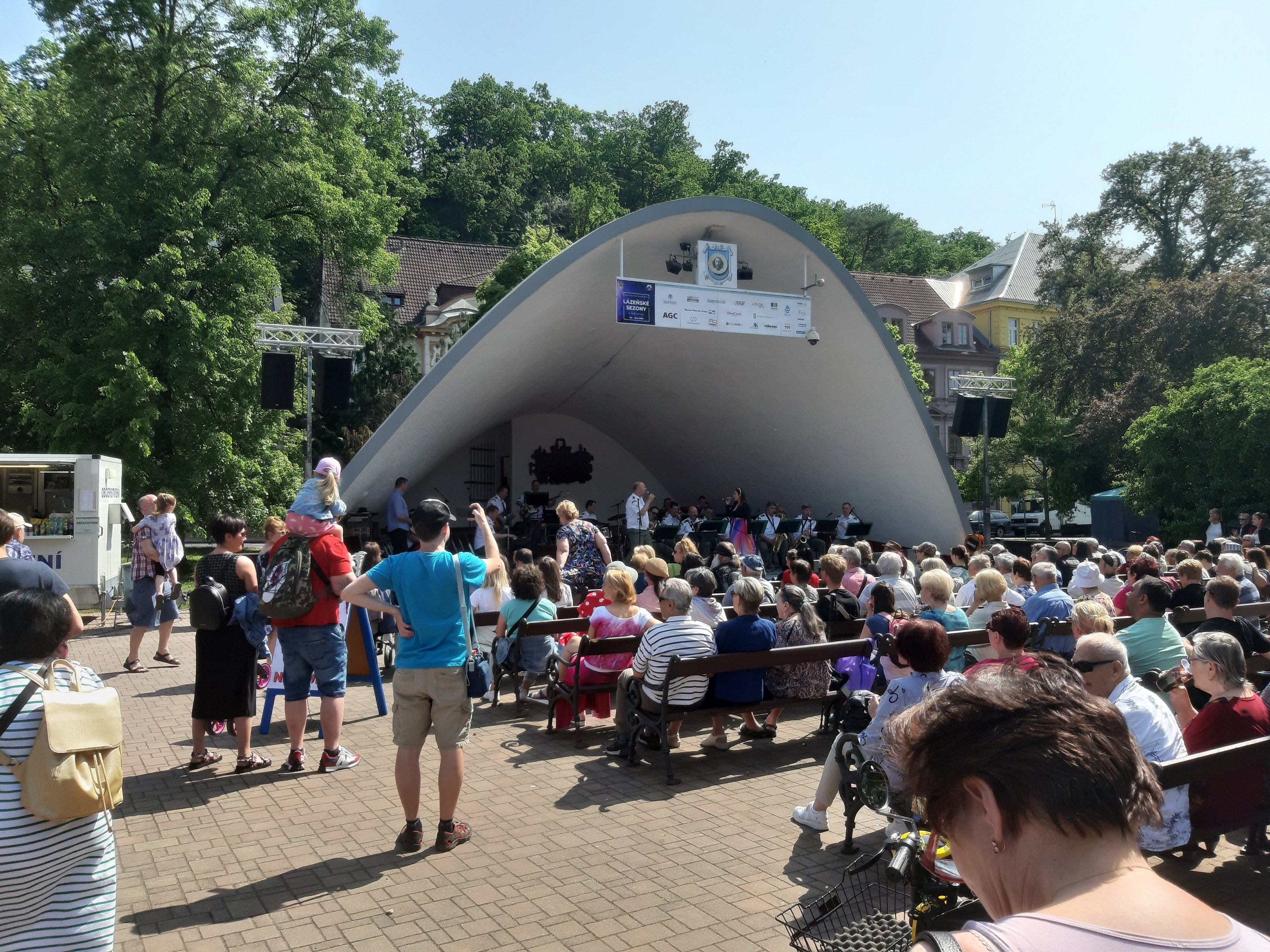 Eine Menschenmenge auf Bänken sitzend und vor einer Bühne an einem Outdoor-Sommerkonzert stehend, mit einem Kameramann, einem Banner, Bäumen, Gebäuden und einem bewölkten Himmel.