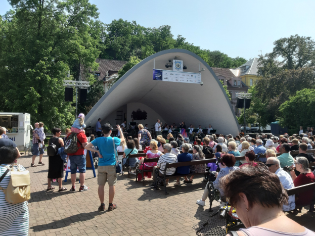 Eine Menschenmenge auf Bänken sitzend und vor einer Bühne an einem Outdoor-Sommerkonzert stehend, mit einem Kameramann, einem Banner, Bäumen, Gebäuden und einem bewölkten Himmel.