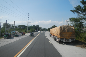 Ein Lastwagen mit einer Plane auf der Ladefläche fährt auf einer Straße, umgeben von Strommasten, Bäumen, Gebäuden und einem bewölkten Himmel, mit einigen Menschen und Fahrzeugen auch auf der Straße.