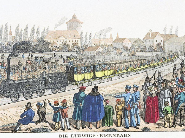 Gruppe von Menschen um einen Zug auf Schienen mit einigen Passagieren im Inneren und anderen außerhalb, mit Gebäuden und Bäumen im Hintergrund und dem Text "Ludwigs-Eisenbahn, die erste Bahnstation in Deutschland" unten.