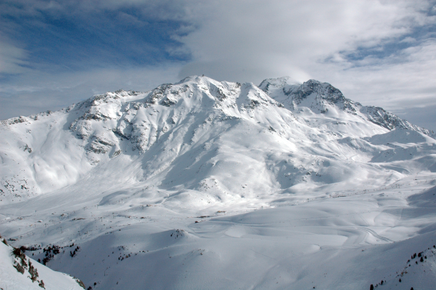 Ein schneebedeckter Berg mit ein paar Skifahrern, die darauf hinunterfahren, umgeben von einer wolkigen Himmel und unberührtem Schnee.