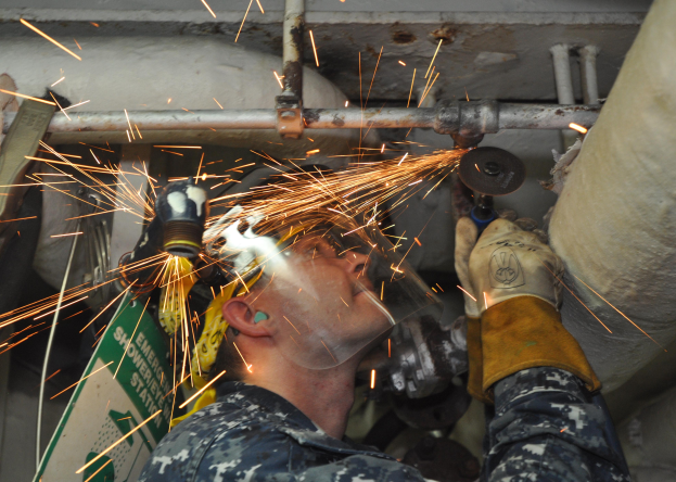 Ein Mann in Helm und Handschuhen bearbeitet ein Metallstück mit einer Schleifmaschine und erzeugt Funken, mit Rohren und anderen Gegenständen im Hintergrund.