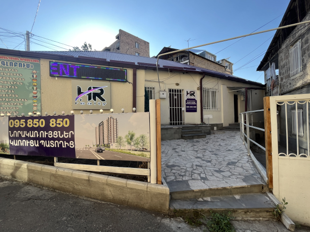 Ein zweistöckiges Gebäude mit einem Metallzaun und Tor, das ein Schild trägt, auf dem "hr Immobilienagentur's Büro zur Miete in Phnom Penh" steht, umgeben von Pflanzen, einem Weg, Pfosten, Drähten, Bäumen und einem bewölkten Himmel.