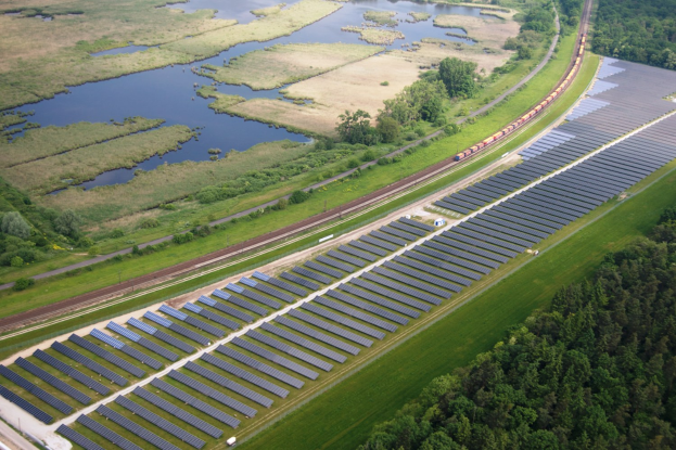 Eine Luftaufnahme eines Solarparks mit zahlreichen Solarpanelen auf einem Feld, umgeben von Bäumen, Gras und Wasser, mit einem Zug, der auf einem Bahngleis in der Nähe fährt.