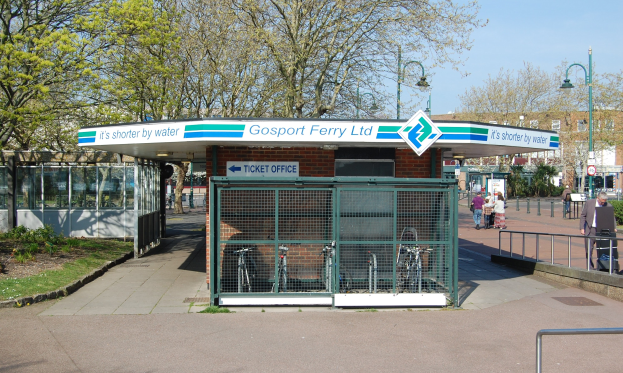 Haltestelle mit einem "Gosport Ferry Ltd."-Schild, Metallzaun, Menschen, Fahrrädern, Bäumen, Straßenlaternen, Schildern, einem Gebäude und einer bewölkten Himmel.