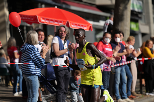 Ein Mann in einem gelben T-Shirt und Laufschuhen sprintet durch einen Marathon, umgeben von einer jubelnden Menge mit einem Schirm und einem Kinderwagen im Hintergrund.