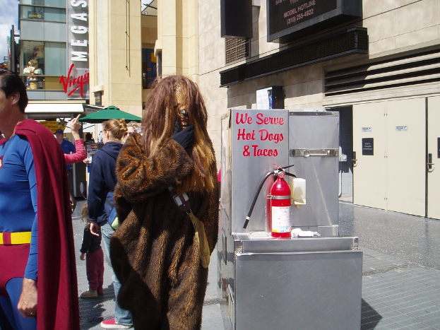 Eine Menge Menschen, einige in Kostümen, versammelte sich vor Gebäuden mit Schildern, einer Box mit Schrift und einem Feuerlöscher.