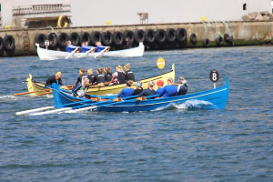 Eine Gruppe von Menschen in einem blauen und gelben Boot auf dem Wasser, die Paddel halten, mit einer Wand aus Reifen und einem Gebäude im Hintergrund, das eine Regatta vermuten lässt.