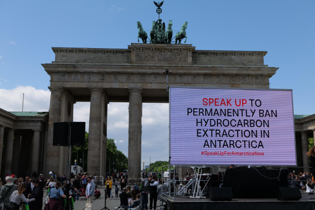 Eine Gruppe von Menschen steht vor dem Brandenburger Tor in Berlin, Deutschland, mit einer Frau auf einer Bühne rechts, Rednern und einem Bildschirm mit der Aufschrift "Sprecht euch aus, um die permanente Verbannung von Kohlenstoffextraktion in der Antarktis zu erreichen". Das Brandenburger Tor zeigt einen Bogen mit Säulen und Statuen, umgeben von Bäumen und einem bewölkten Himmel.