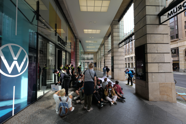 Eine Gruppe von Menschen sitzt und steht vor einem Volkswagen-Geschäft in München, Deutschland, mit Glaswänden, einem Logo und Säulen mit Namensschildern sowie Gebäuden mit Fenstern und Deckenlampen im Hintergrund.