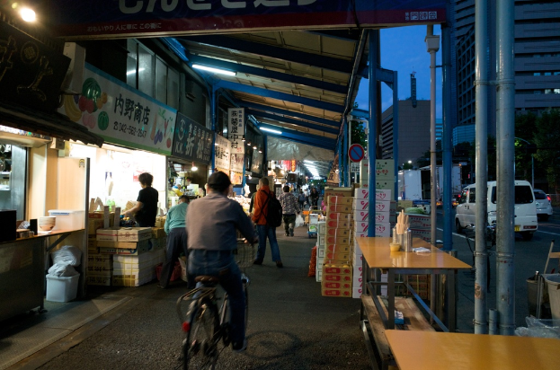 Ein Gehweg mit Geschäften auf der linken Seite und Kisten, einem Tisch, einer Straße mit Autos, Bäumen und einem Gebäude auf der rechten Seite; Personen sind anwesend, eine fährt Fahrrad und eine andere hält eine Tasche, trägt ein oranges Hemd und Jeans.