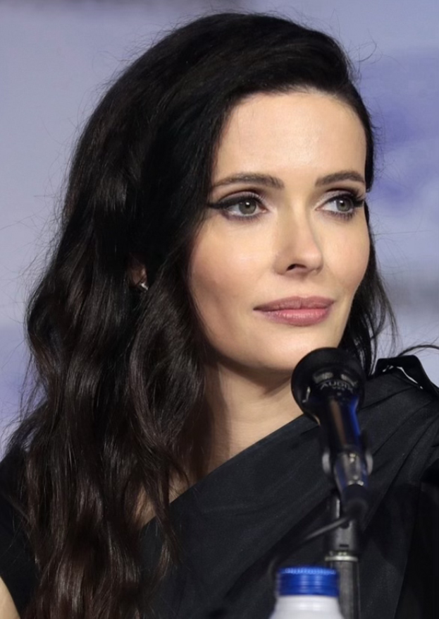 Krysten Ritter auf der Comic-Con in San Diego, in einem schwarzen Kleid, vor einem Mikrofon mit einer Flasche unten, mit einem unscharfen, träumerischen Hintergrund.