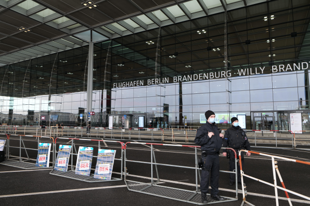 Zwei Polizisten in schwarzen Uniformen stehen vor einem großen Glasgebäude mit Text darauf, mit Absperrungen und Texttafeln im Vordergrund und anderen Menschen, Pfählen, Lichtern im Hintergrund.