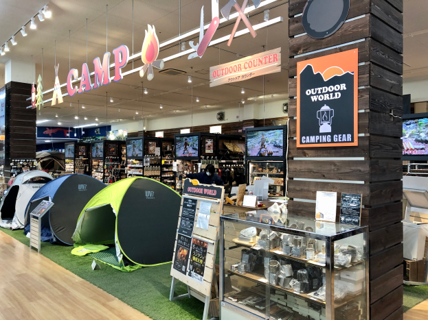 Ein Geschäft voller Campingausrüstung, einschließlich Zelte, Schilder, einer Glasvitrine, Fernsehern, Ständern mit Gegenständen, Deckenleuchten und anderen Objekten.