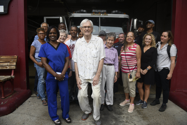 Eine Gruppe von Freiwilligen vor einem Feuerwehrwagen, mit einer Bank links und einer Wand im Hintergrund.