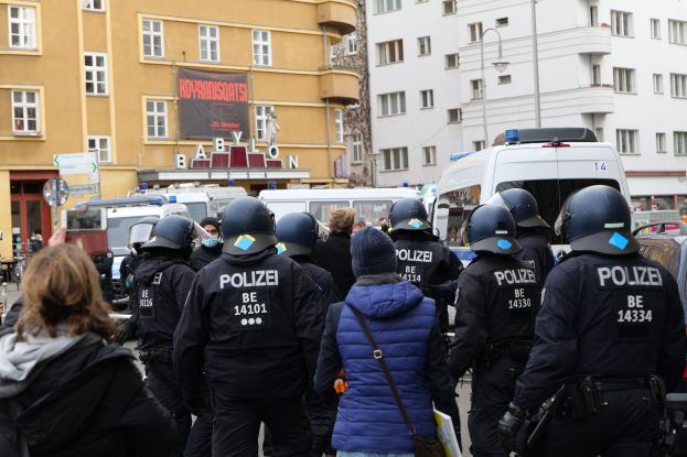 Eine Gruppe von Polizisten steht vor einer Menge von Menschen, die Helme und Jacken tragen, während einer Demonstration in Berlin, Deutschland, mit Fahrzeugen, Gebäuden, Laternenmasten und einem Banner mit Text im Hintergrund und einer Person mit einer Kamera.