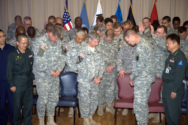 Eine Gruppe von Menschen in militärischer Kleidung steht zwischen roten und blauen Stühlen, mit Flaggen im Hintergrund.