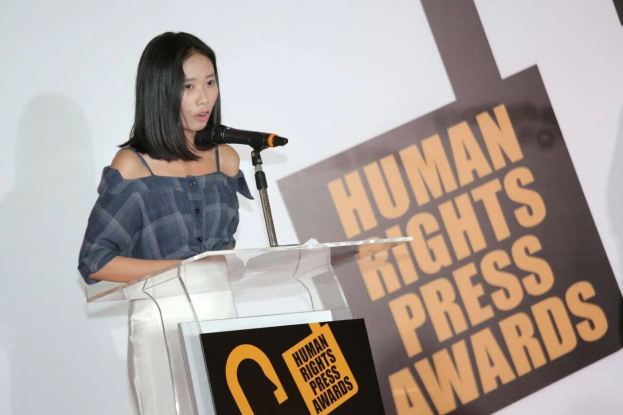 Eine Frau in einem schwarzen Kleid steht an einem Rednerpult mit einem Mikrofon und hält eine Rede bei den Human Rights Press Awards, mit einem ernsten und konzentrierten Gesichtsausdruck.