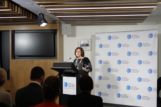 Eine Frau steht auf einer Bühne und spricht in ein Mikrofon, vor ihr sitzt ein Publikum, hinter ihr hängt ein Banner an der Wand, ein Fernseher an der hölzernen Wand und Lampen an der Decke.