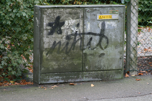 Ein Metallbehälter mit einem Wort und einem Stern, Zahlen und einem Schild auf einem Weg, mit einem Zaun, einem Pfahl, Pflanzen und trockenen Blättern im Hintergrund.