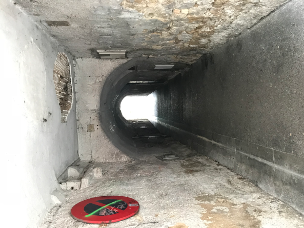 Ein Tunnel mit einem roten und schwarzen "Eintritt Verboten"-Schild auf dem Boden, beleuchtet von Deckenlampen, das darauf hinweist, dass es Teil eines Abwassersystems ist.