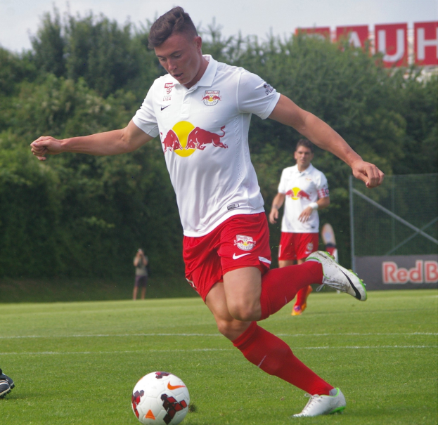 Ein Mann in einem Fussballtrikot schießt einen Ball auf einem grünen Feld mit Bäumen und einem klaren blauen Himmel im Hintergrund, mit einem Schild mit der Aufschrift "RB Leipzig" sichtbar.