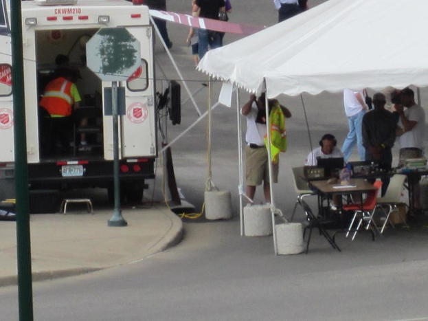 Ein Rettungswagen steht links von einer Straße, während rechts ein Zelt eine Gruppe von Menschen schützt, mit einem Tisch und einem Stuhl in der Nähe.