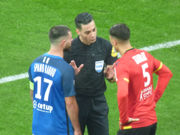 Schiedsrichter in schwarzer Uniform, der mit zwei Männern auf einem Fußballfeld spricht; ein Mann in blauer und roter Kleidung mit Text, beide tragen Brillen.