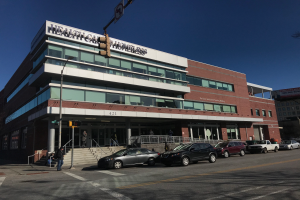 Ein großes Gebäude, das als New York City Health Care Homeless Center identifiziert wurde, mit parkenden Autos davor, Straßenlaternen, Verkehrspfosten, Verkehrszeichen, Menschen auf dem Gehweg, Geländern, Treppen, Bäumen und einem klaren blauen Himmel.