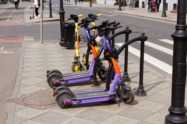 Eine Reihe von Elektro-Scootern, die entlang einer Straße geparkt sind, mit Fußgängern und Fahrrädern auf dem Gehweg, einem Müllcontainer im Vordergrund und Gebäuden, Bäumen und Pflanzen im Hintergrund.