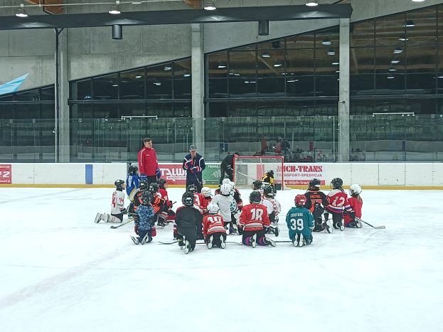 Eine Gruppe von Kindern in Helmen und mit Hockey-Schläger sitzt auf einem Eisring, mit einer Wand aus Glas und Säulen, Deckenleuchten und Texttafeln im Hintergrund.
