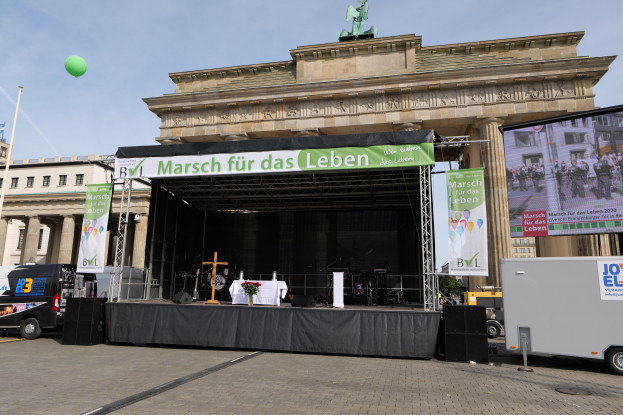 Bühne vor dem Brandenburger Tor in Berlin, Deutschland, mit einem Tisch mit einer Blumenvase, einem Kreuzsymbol, Transparenten mit Text, Lautsprechern und anderen Gegenständen, sowie Fahrzeugen auf der Straße und Gebäuden im Hintergrund.