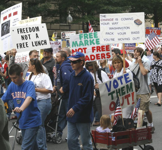 Eine Gruppe von Menschen, die auf einer Straße mit Brettern gehen, mit Kindern in einem Wagen vorne und einem Baum, einem Lichtmast und einem Gebäude im Hintergrund.