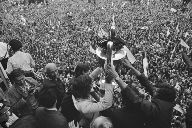 Schwarzes Bild einer Gruppe von Menschen, die mit einem Pokal in der Luft feiern, vor einer großen Menge, einige halten Kameras und Fahnen.