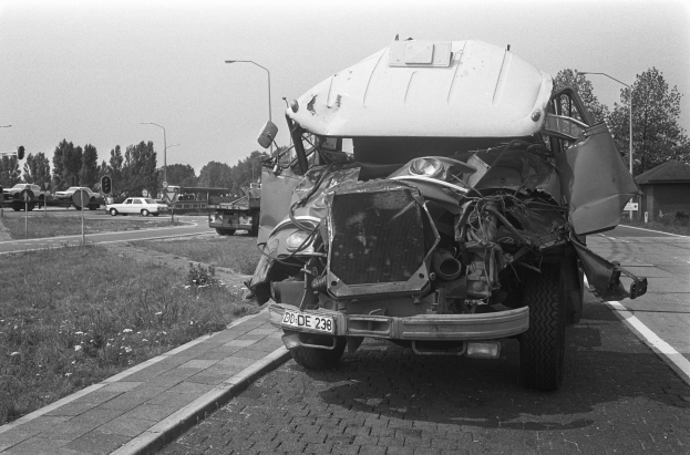Ein Schwarz-Weiß-Foto eines schwer beschädigten Autos am Straßenrand mit eingedellter Motorhaube, zersplitterten Fenstern und anderen Fahrzeugen, Laternenpfählen, Bäumen und einem Haus im Hintergrund.
