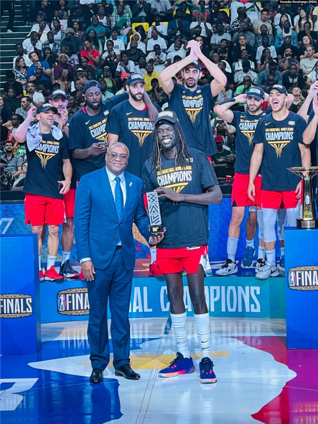 Gruppe von Männern, die auf einem NBA-Finals-Podium feiern, zwei halten Trophäen, zusätzliche Trophäen auf Podesten und Zuschauer im Hintergrund sitzend.