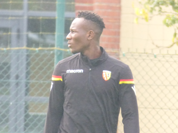 Ein Mann In Einer Schwarzen Jacke Steht Auf Einem Fussballfeld, Umgeben Von Einer Umzäunung Und Einem Baum, Scheinbar Fokussiert Während Einer Trainingseinheit.