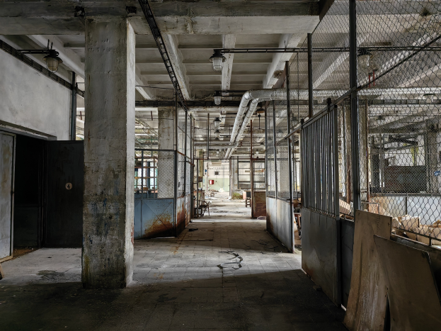 Leerer Raum mit großen Fenstern, Säulen, einer Tür, Deckenleuchten, Metallstangen, einem Maschendrahtzaun, verstreuten Gegenständen auf dem Boden und einer Wand, die eine verlassene Fabrik mit einem vernachlässigten Erscheinungsbild zeigt.