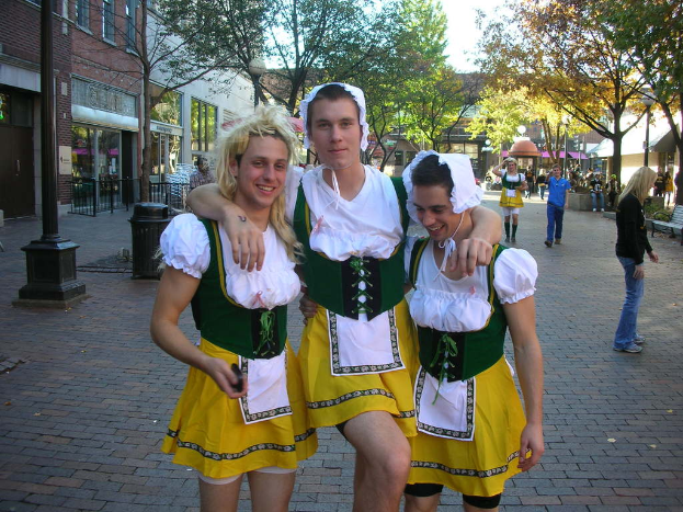 Drei Männer in weißen und gelben Kostümen stehen lachend vor einer belebten Straßenszene mit Menschen, Gebäuden, Bäumen, einer Bank, einem Müllcontainer und einem Geländer.