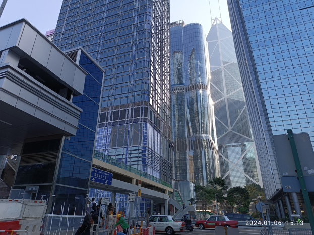 Eine belebte Stadtstraße mit hohen Wolkenkratzern, fahrenden Fahrzeugen, Fußgängern auf dem Gehweg, Absperrpoller, Straßenschilder, Bäumen und einem klaren blauen Himmel.